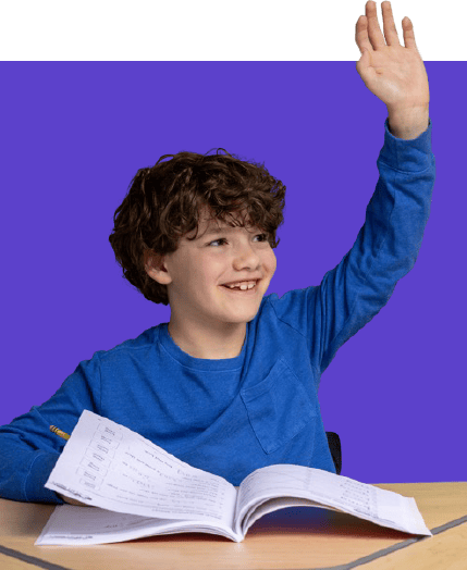 Smiling boy in a blue shirt raises his hand while looking to the side, seated at a desk with an open workbook.